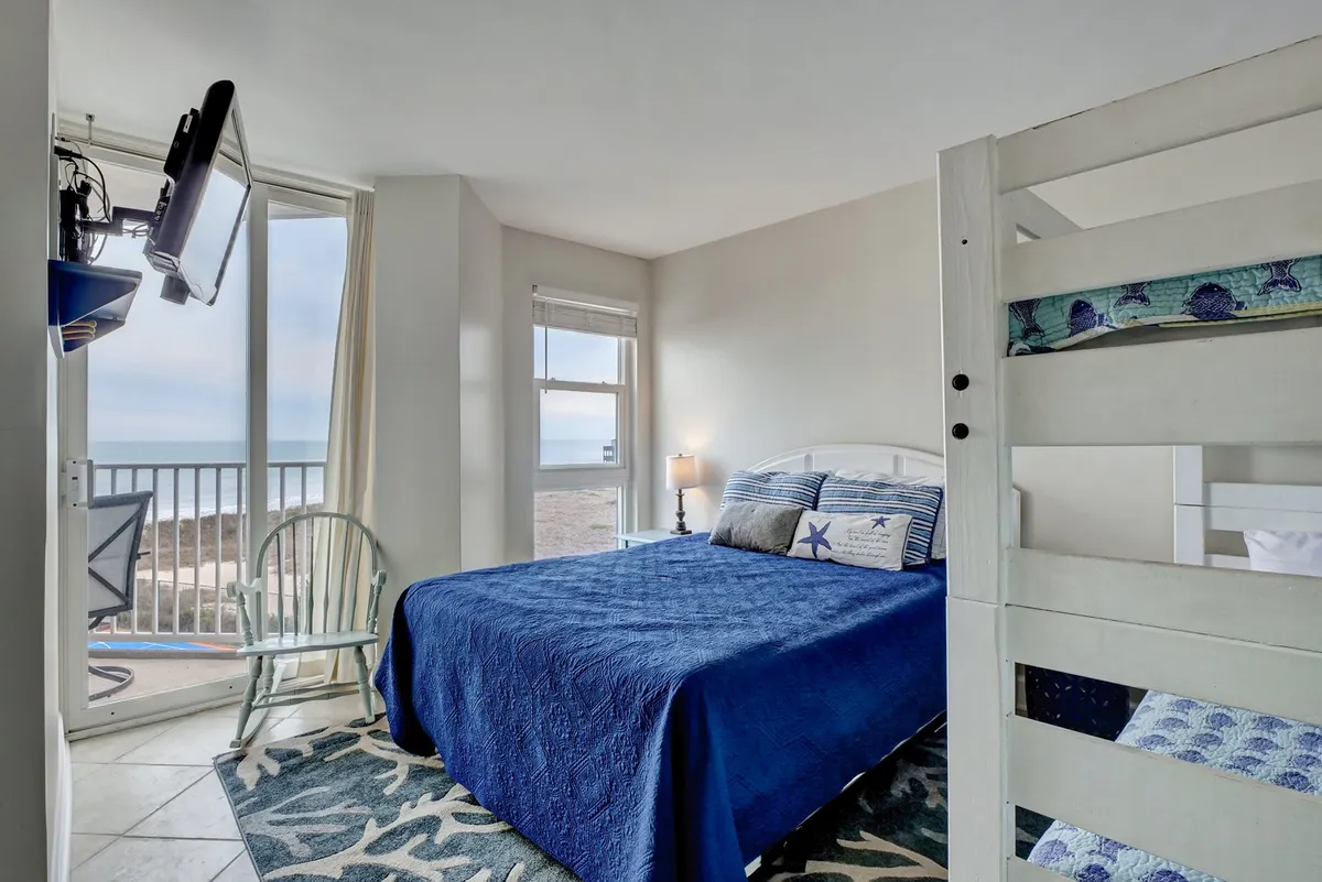 A tranquil, family-friendly bedroom featuring a plush blue bed, a bunk bed suitable for children or friends, large windows overlooking the beach, and a mounted television. Decorative accents add a welcoming touch.