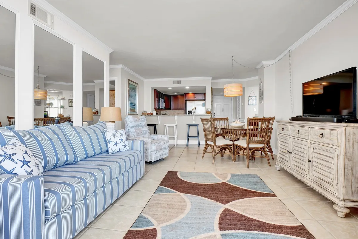This image features a spacious, open-concept living area with a striped blue sofa, a wooden dining table, a television, and a cozy area rug. The room exudes comfort and is clearly set up for relaxation, entertaining, and family meals, with a casual and welcoming atmosphere.