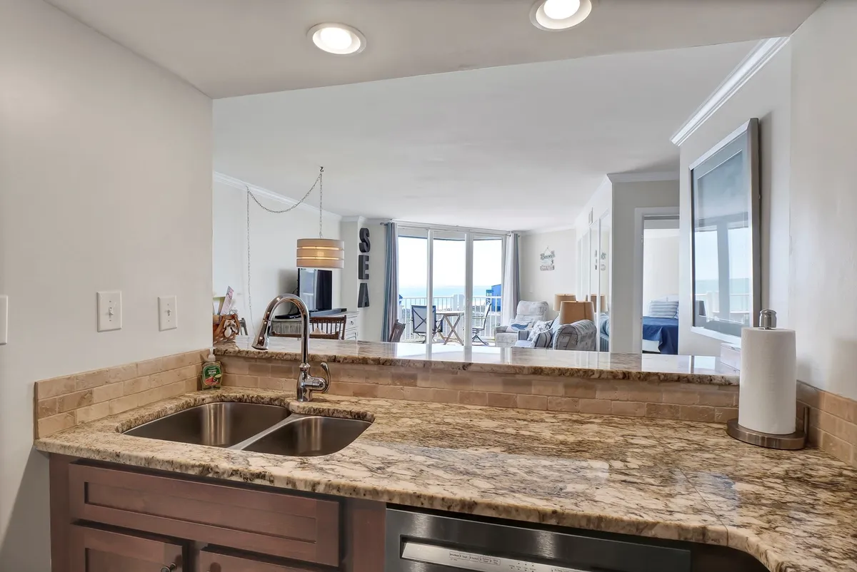 This image features a kitchen area with a granite countertop and sink, looking out into a living space with ocean views. Decorative elements like a 'SEA' sign reinforce the coastal theme, and the overall mood is relaxed and inviting, perfect for entertaining or enjoying a morning coffee.