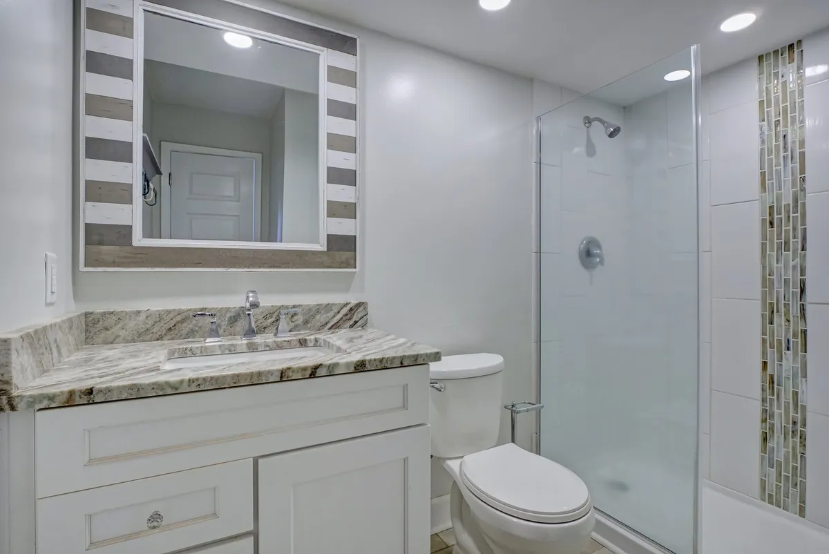 A fresh and inviting bathroom interior, featuring a granite countertop, a large striped mirror, and a glass shower enclosure. Subtle decor suggests a spa-like environment, perfect for relaxing after a beach day.