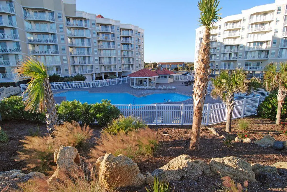A sparkling swimming pool surrounded by palm trees and resort buildings, with inviting lounge chairs arranged around the pool. Groups of people are seen swimming, sunbathing, and socializing, evoking a cheerful, communal resort atmosphere.