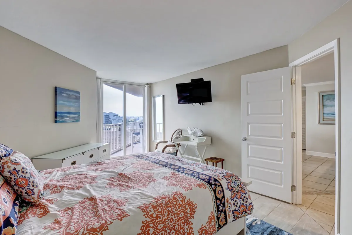 This image showcases a bedroom with colorful bedding, large windows that allow natural light to flood the space, a wall-mounted TV, and a desk and chair setup. The view through the windows suggests a tranquil setting, ideal for relaxation.