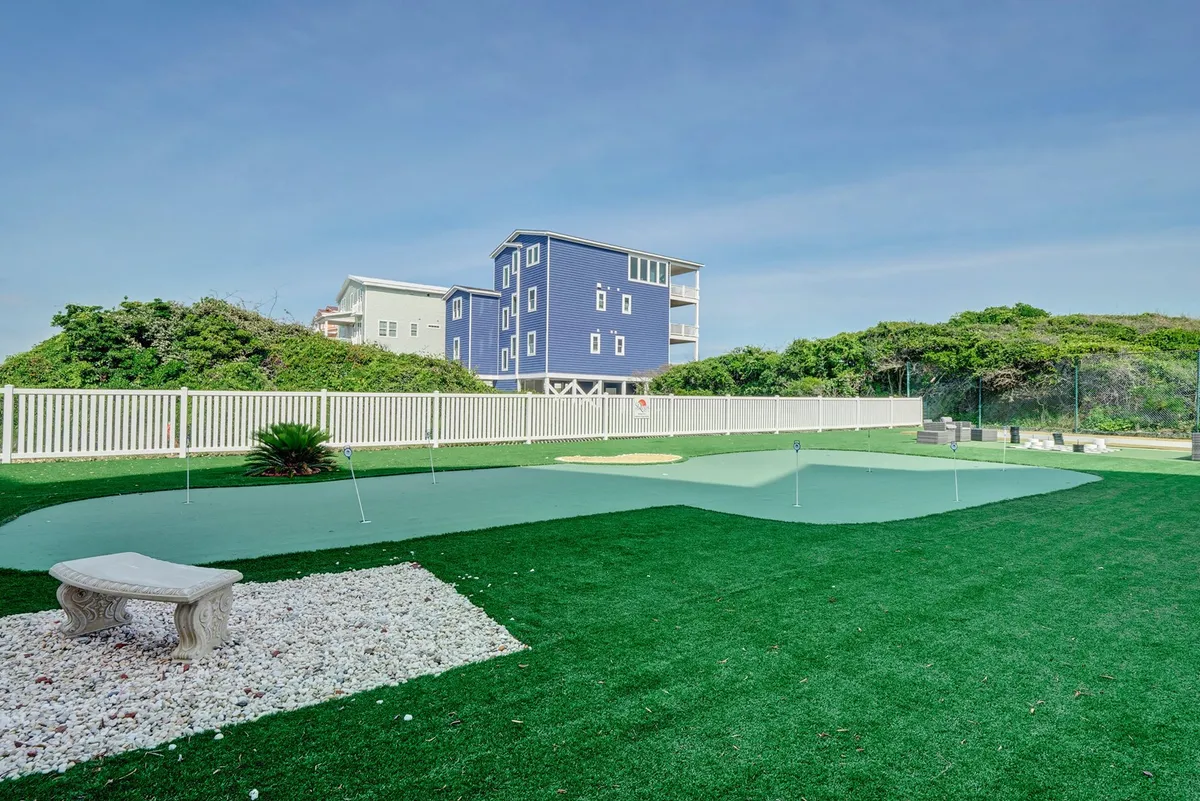 This exterior image showcases an artificial putting green with a stone bench and lush greenery surrounding the area, with a blue house in the background. The scene is inviting and tranquil, suggesting a space where guests can both relax and engage in light outdoor activities.