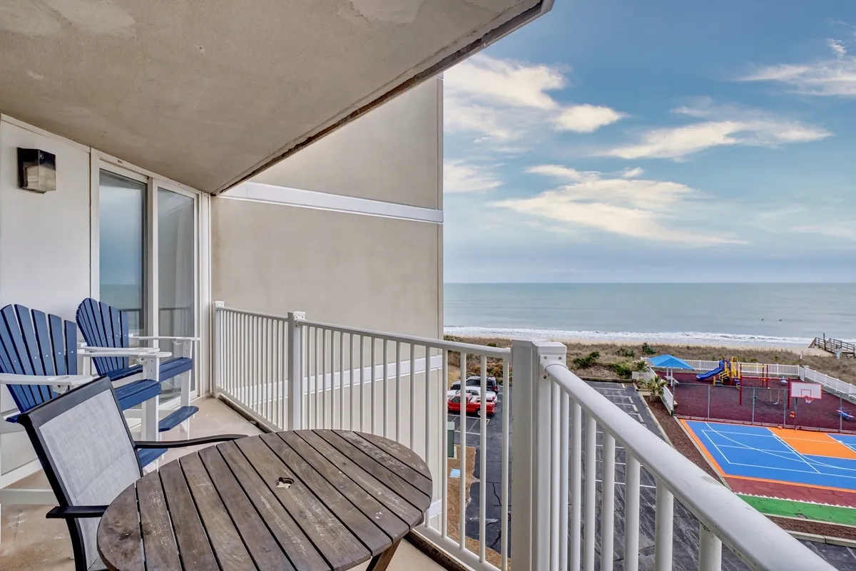 This exterior image features a spacious balcony with comfortable seating overlooking the ocean and a colorful sports court below. It captures both relaxation and active recreation opportunities, as you can see the tranquil water views as well as the area where sports like basketball and pickleball take place. The sunrise light and the inviting balcony setting evoke the perfect family retreat by the sea.