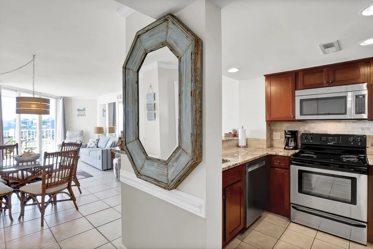 This interior shot captures a dining table, a mirrored wall, visible kitchen appliances, and a spacious living area illuminated by large windows. The scene is warm and inviting, suggesting social gatherings and moments of relaxation, with the windows hinting at potential ocean views.