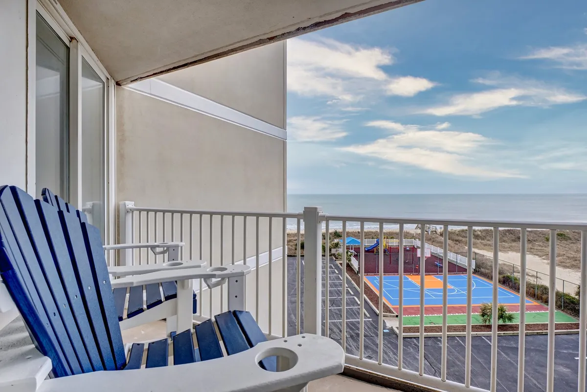 A private balcony furnished with blue and white Adirondack chairs, offering a sweeping ocean view. Below, a colorful sports court is visible, suggesting opportunities for recreation right at the property. The balcony setting feels perfect for social gatherings, relaxing sunrises, or taking in the sights of friends playing sports below.